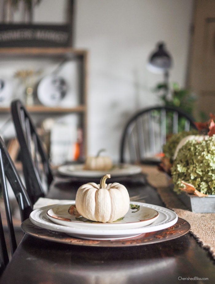 A simple Farmhouse Fall Tablescape with a hydrangea and pumpkin centerpiece, burlap table runner, and fall leaves to accent the simple place settings. 
